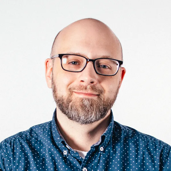 A man with a bald head, glasses, and a beard is smiling slightly. He is wearing a blue button-up shirt with small white patterns, and the background is a plain light color.